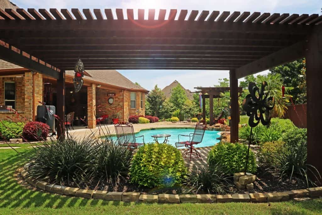 A backyard with a pergola, metal chairs around a small table, landscaped plants, and a swimming pool near a brick house on a sunny day—perfect for outdoor living photos featuring Ellis Landscape Services work.