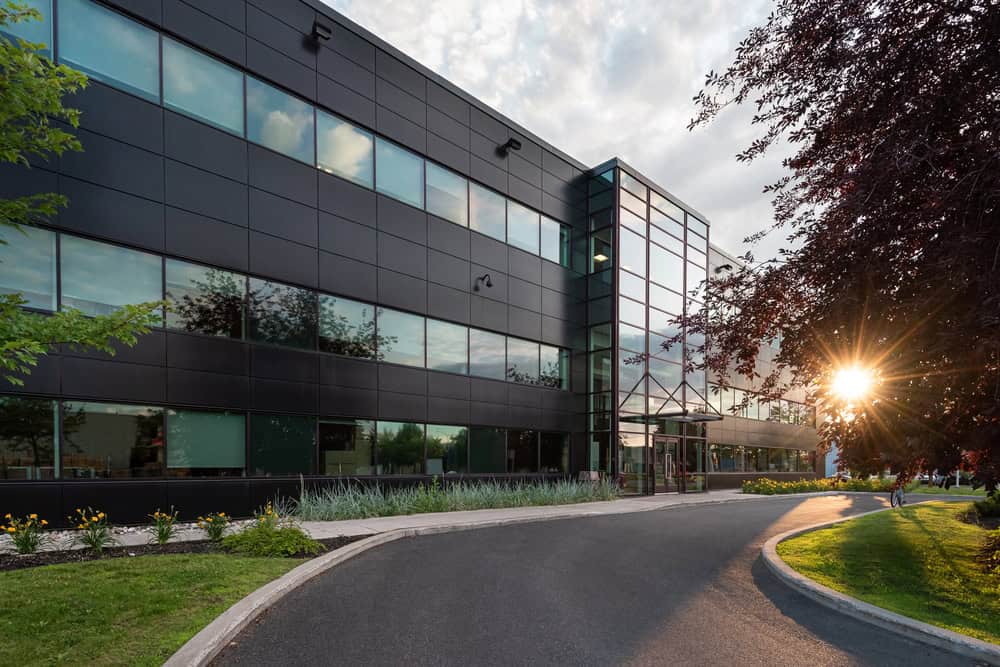 Modern office building with large windows and dark exterior panels, surrounded by professional grounds maintenance and commercial landscaping in Tarrant County, at sunset with sunlight filtering through trees. Modern office building with large windows and dark exterior panels, surrounded by professional grounds maintenance and commercial landscaping in Tarrant County, at sunset with sunlight filtering through trees.