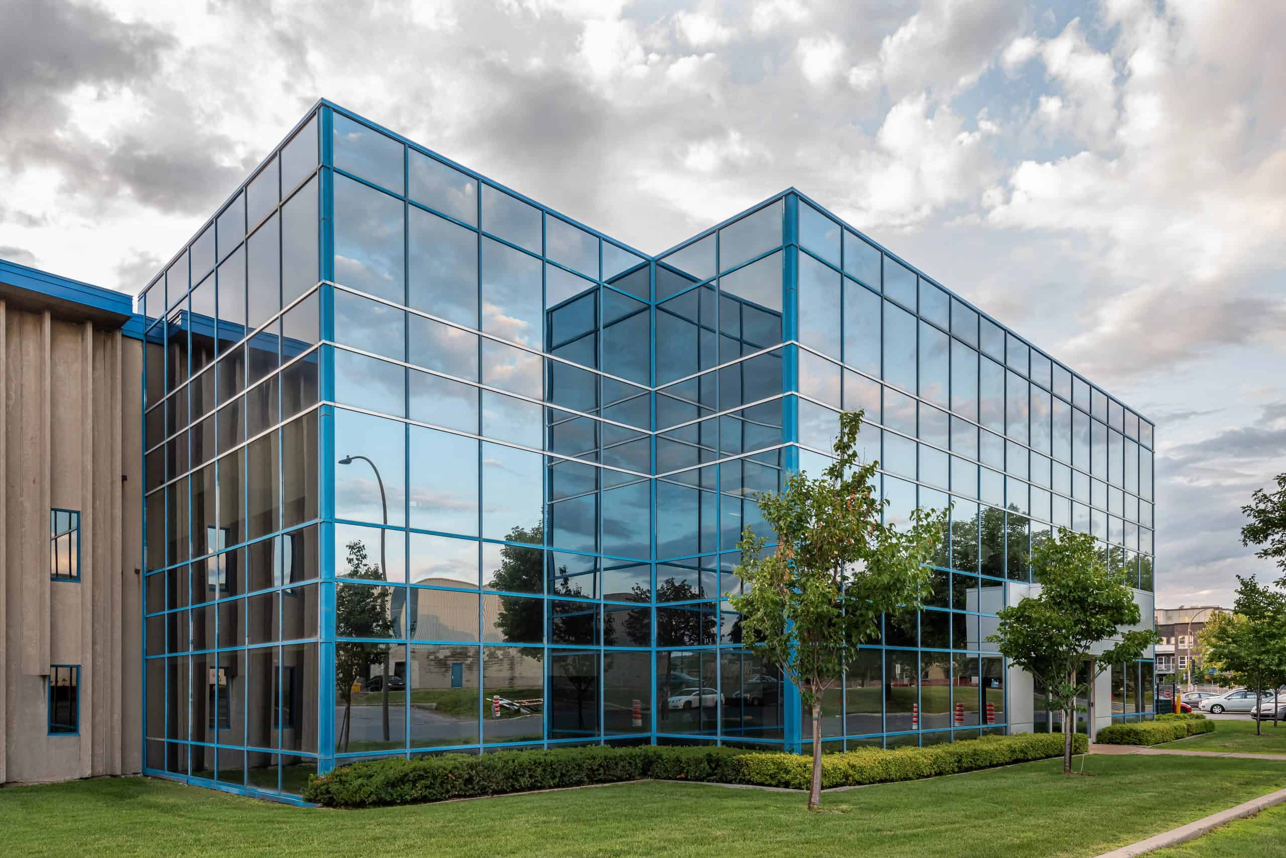 Modern commercial building with blue-tinted glass windows reflecting the sky and surroundings, set on a grassy lawn with small trees and enhanced by professional grounds maintenance.