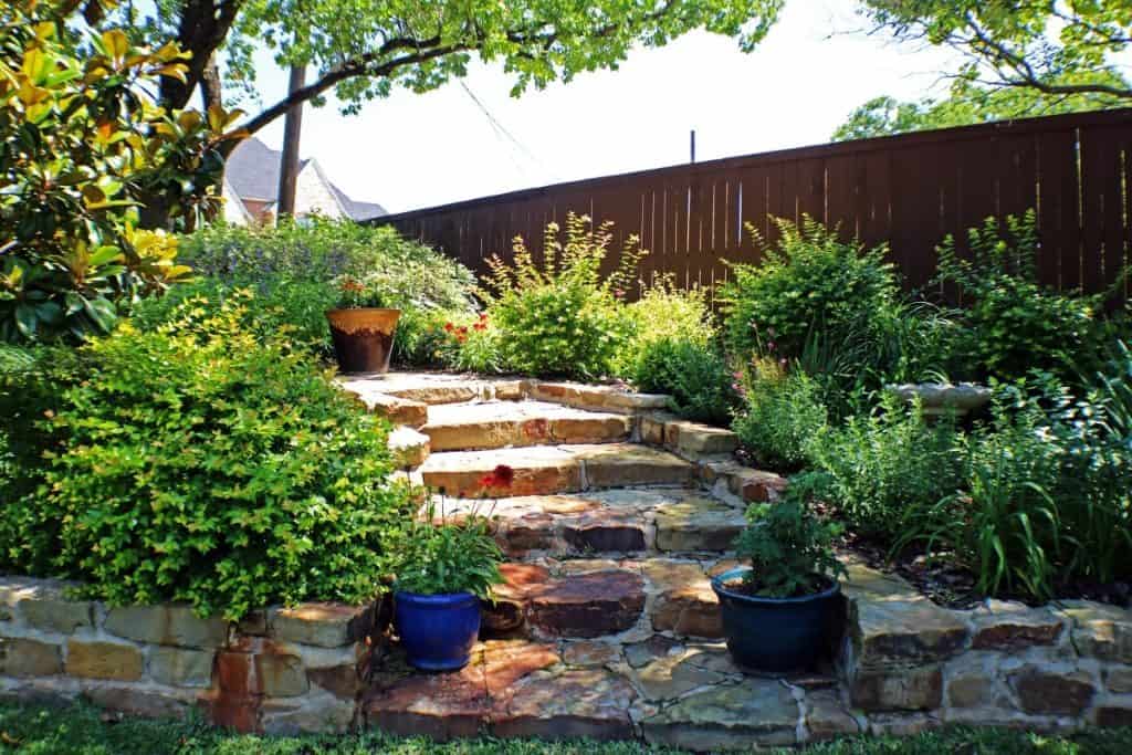 Stone steps lead up through a lush garden with green shrubs, potted plants, and a wooden fence—an inviting example of hardscaping Tarrant County homeowners love. Stone steps lead up through a lush garden with green shrubs, potted plants, and a wooden fence—an inviting example of hardscaping Tarrant County homeowners love.