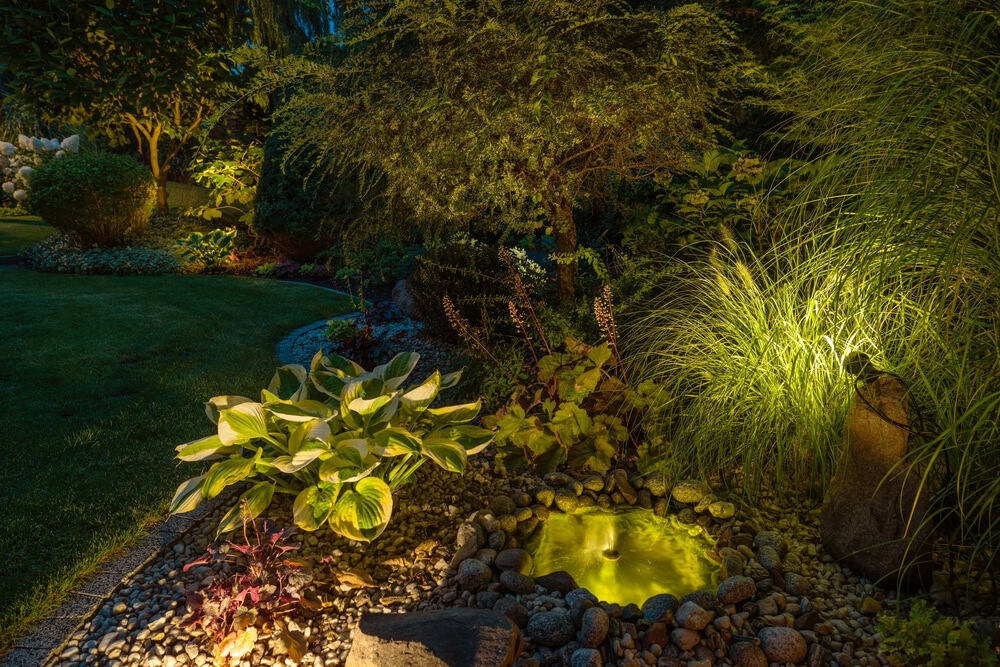 A small illuminated garden fountain surrounded by rocks, ornamental grasses, and various plants at night, enhanced by elegant accent lighting Tarrant County to create a magical evening atmosphere.