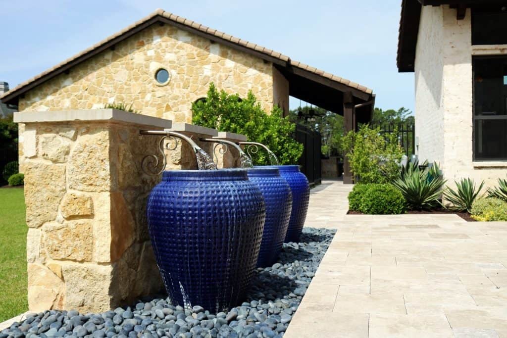 Three large blue ceramic water fountains stand on dark pebbles beside a stone patio and building, creating stunning outdoor fountains Tarrant County homeowners love in a beautifully landscaped backyard.