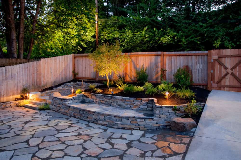 A backyard in Tarrant County featuring hardscaping with a stone patio, raised garden bed, small shrubs, wooden fence, and inviting outdoor lighting at dusk—perfect for your outdoor living dreams.