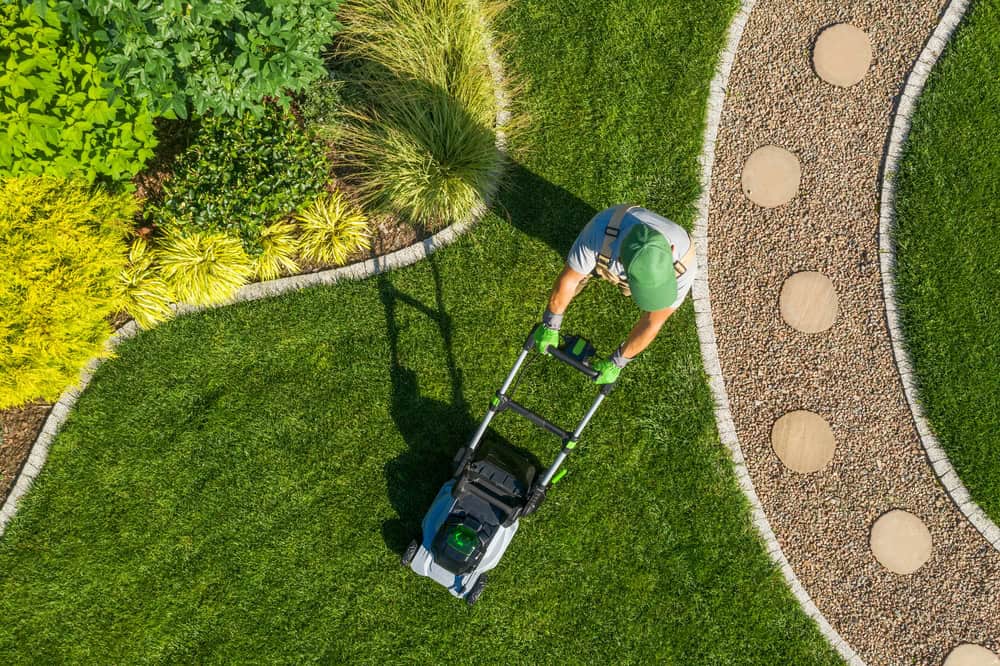 Person mowing a well-manicured lawn with a push mower, surrounded by landscaped plants and a decorative stone pathway, showcasing expert lawn care Tarrant County. Person mowing a well-manicured lawn with a push mower, surrounded by landscaped plants and a decorative stone pathway, showcasing expert lawn care Tarrant County.