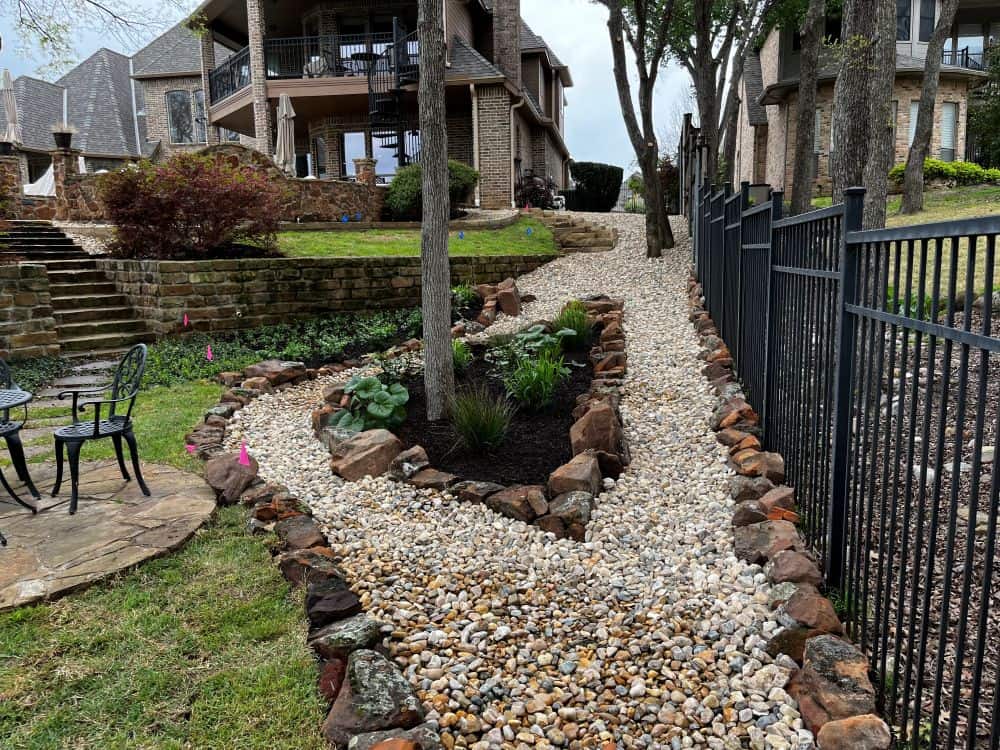 A landscaped backyard designed with expert Landscape Services features a stone-bordered gravel pathway, a small garden bed, metal fence, and patio chairs, all set against a backdrop of multi-story houses.