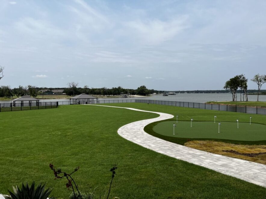 A curved sidewalk, crafted by expert Landscape Services, borders a manicured lawn and putting green near the waterfront, with docks and trees in the background under a partly cloudy sky.