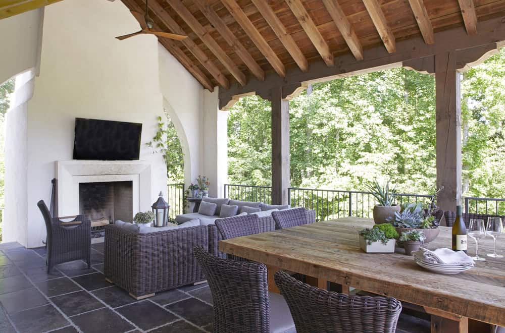 Covered outdoor patio with a wooden ceiling, wicker seating around a fireplace and TV, and a wooden dining table set with plants and wine glasses, showcasing modern hardscape trends in North Texas and overlooking green trees.