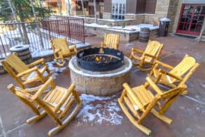 Six wooden rocking chairs circle a metal fire pit on a stone patio with patches of snow—an inviting winter retreat that highlights the charm of outdoor living spaces in North Texas.