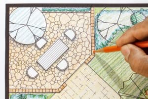 A hand uses an orange pencil to draw on a colored winter landscape planning design plan featuring a patio with a table, four chairs, and surrounding greenery—perfect for Texas outdoor living.