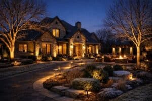 Large stone house with lit windows and landscaped garden at dusk, featuring LED outdoor lighting North Texas style. An inviting outdoor seating area with a firepit and pathway lights beautifully illuminate the trees and shrubs.