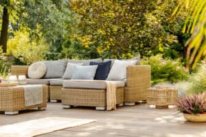 Wicker outdoor sofa with gray and blue cushions on a wooden deck, perfect for summer backyard design and outdoor entertaining spaces, surrounded by plants and trees in a sunlit garden.