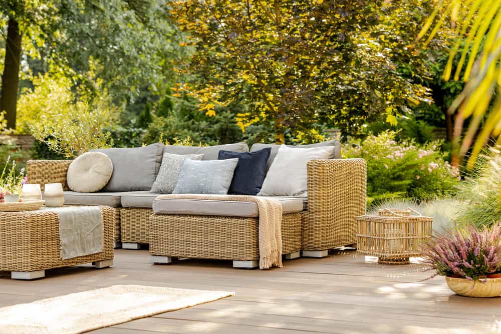 Wicker outdoor sofa with gray and blue cushions on a wooden deck, perfect for summer backyard design and outdoor entertaining spaces, surrounded by plants and trees in a sunlit garden.